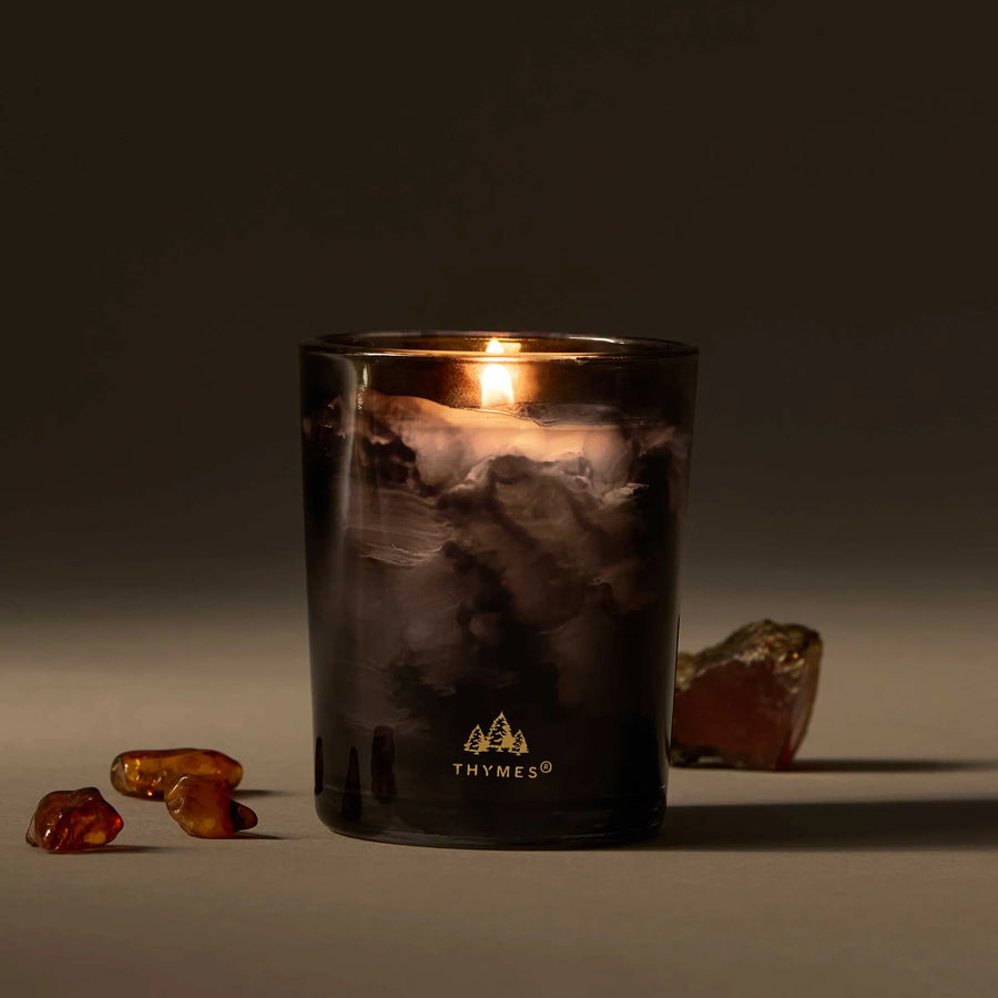 A lit candle in a black and grey smokey glass sits against a grey-brown background, with amber colored crystals around it.