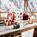 Three mini tin candles sitting on a wooden picnic table, with the accompanying box behind them. The candles are surrounded by other holiday elements such as fruits and a red dutch oven, with a snowy mountain landscape behind them.