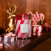 A large, red and white striped glass jar candle sits on a wooden table, with a mini tin candle with red and white stripes to its right. There are candy canes and peppermints around the two candles, and a gold deer figurine in the background.