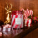 A large, red and white striped glass jar candle sits on a wooden table, with a mini tin candle with red and white stripes to its right. There are candy canes and peppermints around the two candles, and a gold deer figurine in the background.