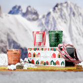 A box with cartoon holiday houses sits on a toboggan with three votive candles sitting around the scene. In the background, snow and mountains can be seen out of focus.