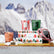 A box with cartoon holiday houses sits on a toboggan with three votive candles sitting around the scene. In the background, snow and mountains can be seen out of focus.