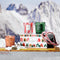 A box with cartoon holiday houses sits on a toboggan with three votive candles sitting around the scene. In the background, snow and mountains can be seen out of focus.
