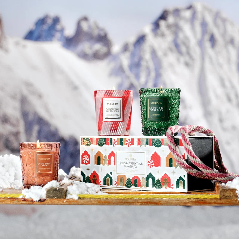 A box with cartoon holiday houses sits on a toboggan with three votive candles sitting around the scene. In the background, snow and mountains can be seen out of focus.