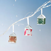 Three boxed holiday candles hang on a white flocked branch, in front of a light blue background.
