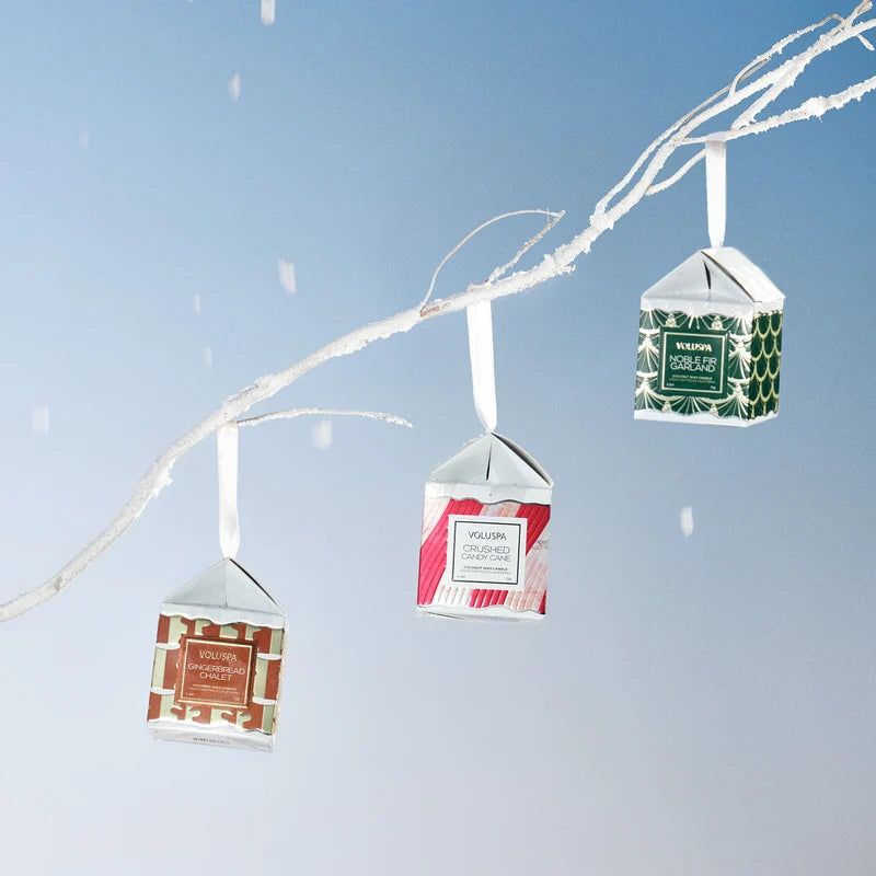 Three boxed holiday candles hang on a white flocked branch, in front of a light blue background.