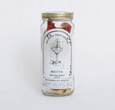 A clear glass jar with a gold metal screw-on lid, containing dried fruit pieces, with a white label reading 'Botanical Craft Cocktails Watermelon Mojito'. The jar is photographed against a white background.
